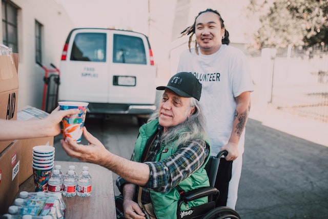 An elderly man in a wheelchair receives food at a soup kitchen. A young man wearing a volunteer shirt pushes the older man's wheelchair. A white van is in the background.