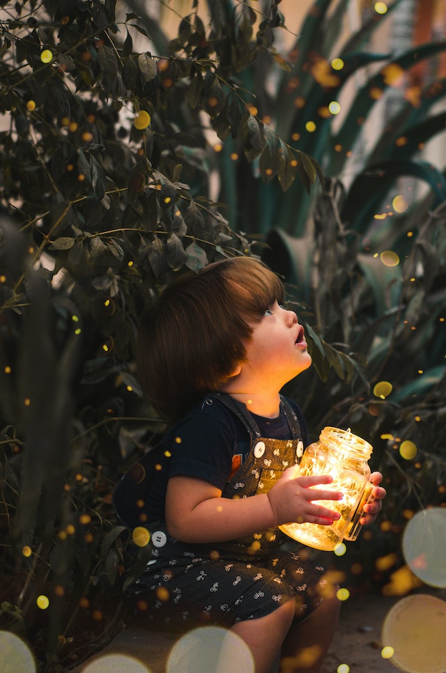 A small child holds a clear glass jar holding fairy lights, looking up in wonder.