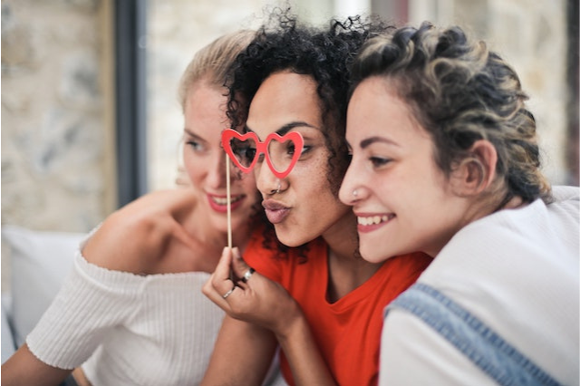 A trio of friends take a silly picture together. A black woman with curly hair stands in the middle, holding cardboard heart-shaped glasses on a stick to her eyes. A white woman stands on either side of her, smiling for the camera.