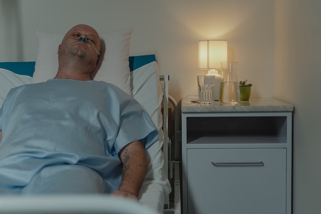 A bald man lies in his hospital bed. The room is dimly lit.