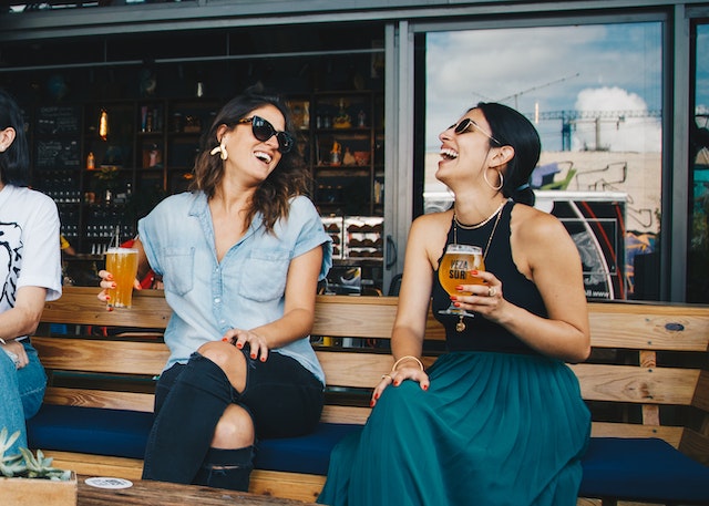 Two young women sit together, laughing, with beer in hand at a local taproom.