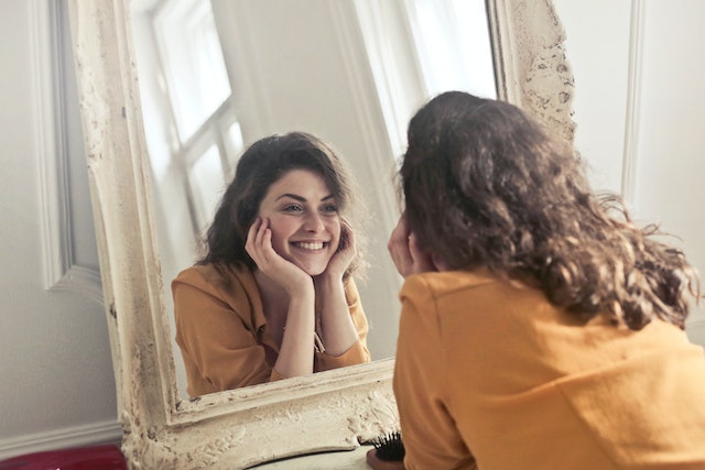 A young white woman with brown hair smiles at her reflection in a mirror.