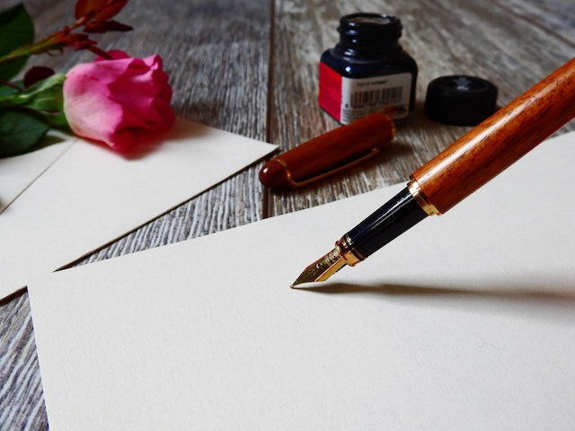 A fountain pen is poised above a blank piece of stationery. An inkwell and pink rose sit in the background.