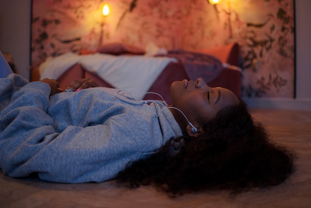 A young black woman lies peacefully in a dim room with her earbuds in and candles lit behind her.